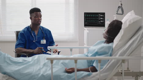 A-black-male-doctor-is-talking-to-a-black-female-patient-lying-on-a-hospital-bed-and-connected-to-an-acid-supply.-The-attending-physician-discusses-with-the-patient-in-the-hospital-the-treatment